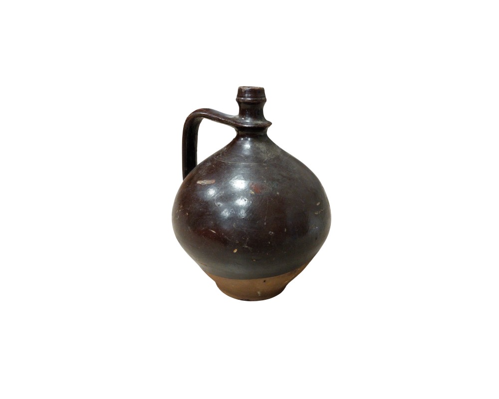 A ceramic jug with a globular body and a glossy glazed finish in dark brown tones, complemented by a visible earthen base and a side handle that connects to the narrow neck; available for hire for events, advertising, films and TV series