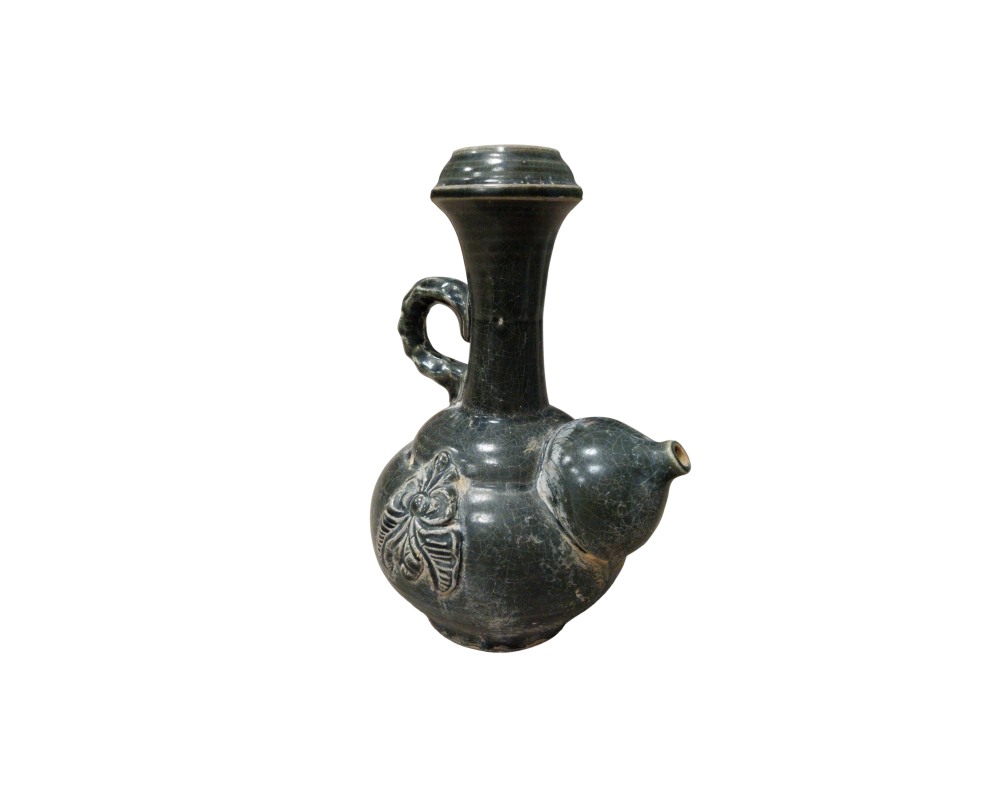 jarra de cerámica con un distintivo acabado esmaltado en tonos oscuros y efecto craquelado, caracterizada por un cuerpo globular que incluye un relieve floral modelado, un asa lateral curvada, un cuello alto ensanchado en la parte superior y un apéndice vertedor lateral alquiler para eventos, publicidad, peliculas y series