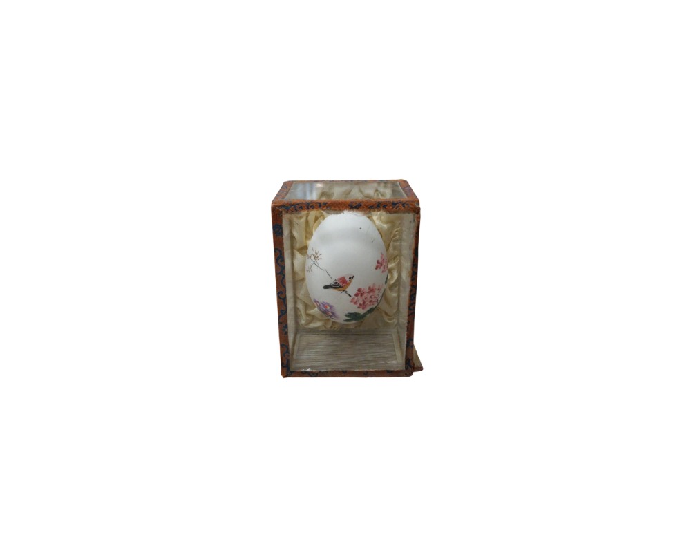 A small, egg-shaped object in shades of white, delicately hand-painted with floral motifs and a bird perched on a branch. It is displayed inside a display case or upright box with a structure covered in patterned fabric and an interior lined with soft fabric; available for hire for events, advertising, films and TV series.