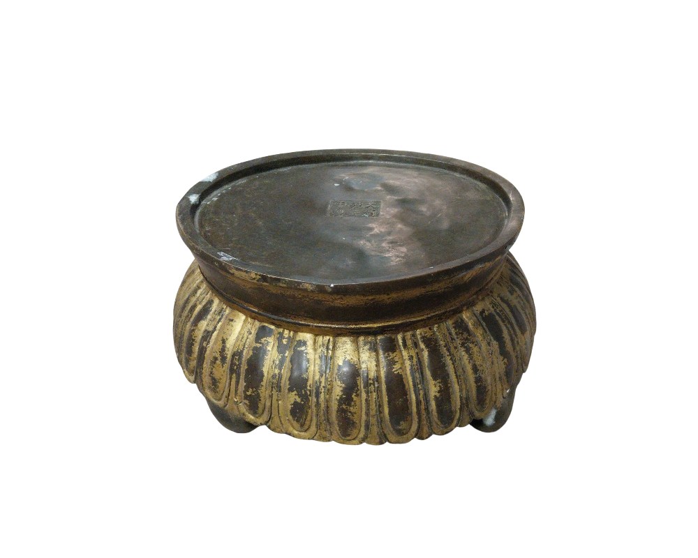 Objeto metálico de forma circular, caracterizado por una estructura baja que descansa sobre pequeñas patas. Su diseño presenta un cuerpo acanalado con un acabado parcialmente dorado que contrasta con el tono oscuro del metal, y en su superficie superior se observa una marca de sello grabada. Es una pieza de manufactura artesanal con una estética clásica, que puede utilizarse como base decorativa para elevar objetos o como un recipiente ceremonial tradicional.