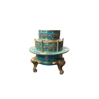 An ornamental container arranged in tiers, characterised by a cloisonné-style enamel finish in shades of turquoise blue and multicoloured floral details, metal panels with figurative reliefs, gilded edges and a lower section supported by three legs sculpted in the shape of an elephant’s head and trunk; available for hire for events, advertising, films and TV series