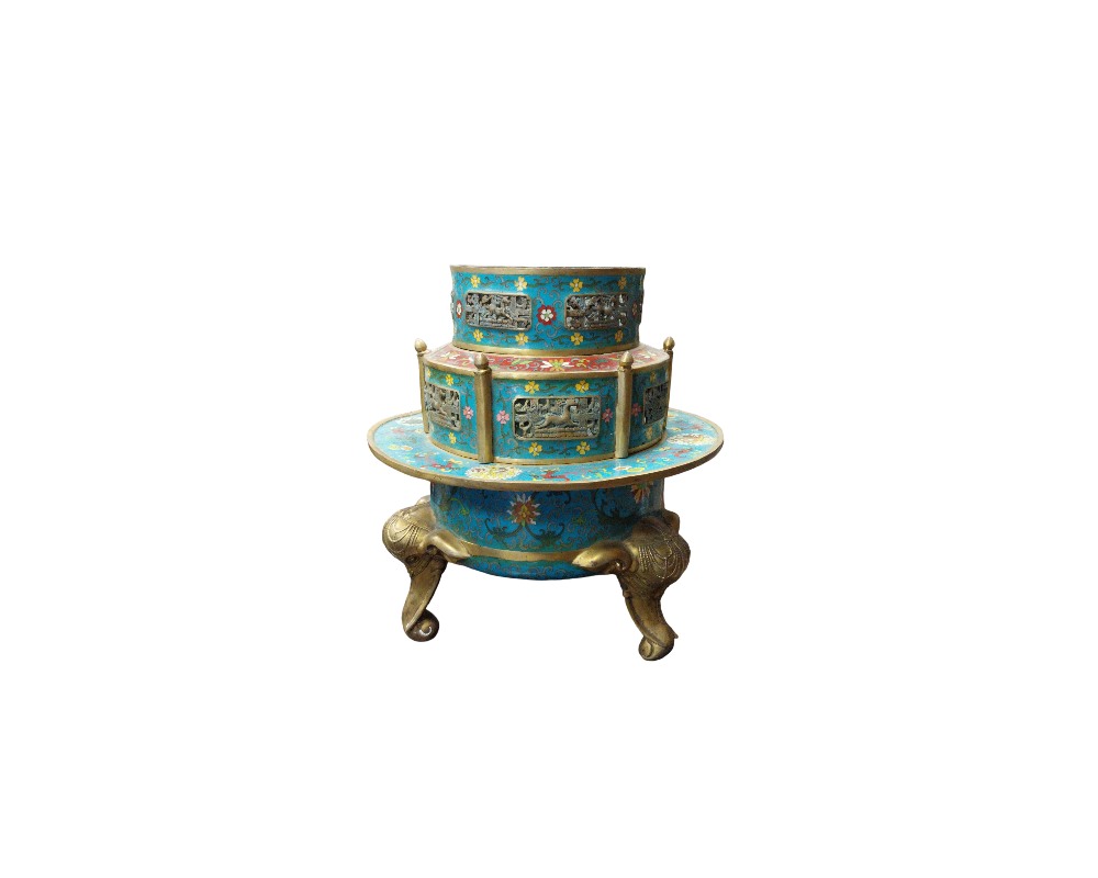 An ornamental container arranged in tiers, characterised by a cloisonné-style enamel finish in shades of turquoise blue and multicoloured floral details, metal panels with figurative reliefs, gilded edges and a lower section supported by three legs sculpted in the shape of an elephant’s head and trunk; available for hire for events, advertising, films and TV series