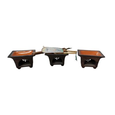 The set comprises three small ceramic stands or braziers, square in design, with openings at the bottom and a rustic finish in dark tones with orange interiors. Available for hire for events, advertising, films and TV series.