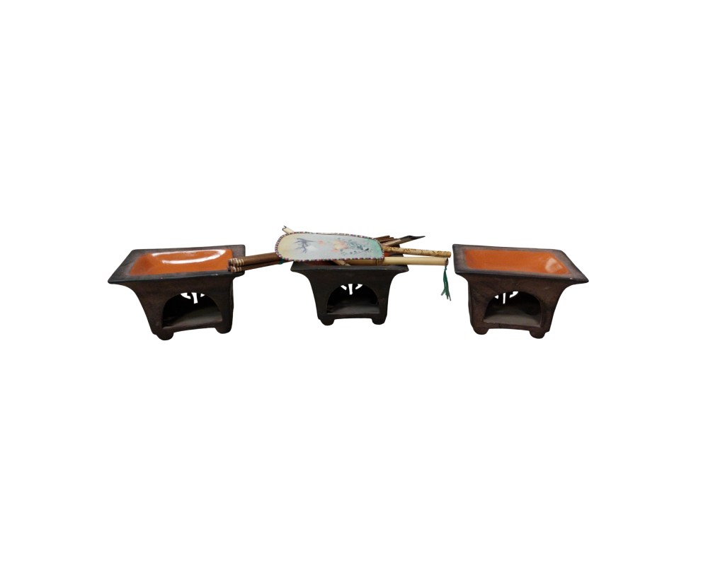 The set comprises three small ceramic stands or braziers, square in design, with openings at the bottom and a rustic finish in dark tones with orange interiors. Available for hire for events, advertising, films and TV series.