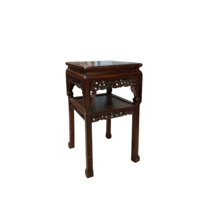 A wooden side table with a dark finish and visible grain, characterised by a square design, a top with moulded edges, a lower shelf and skirting decorated with fine openwork carvings of traditional motifs; available for hire for events, advertising, films and TV series