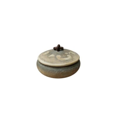 pequeño recipiente circular con textura rústica, caracterizado por una base redondeada, una tapa superior plana y ornamentada con motivos claros en relieve, y un pequeño tirador central de diseño singular alquiler para eventos, publicidad, peliculas y series