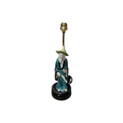 lámpara de sobremesa estructurada a partir de una estatuilla de cerámica esmaltada que representa a un personaje tradicional anciano con túnica azulada y sombrero cónico, montada sobre una base circular oscura y equipada con un vástago y portalámparas metálico superior alquiler para eventos, publicidad, peliculas y series