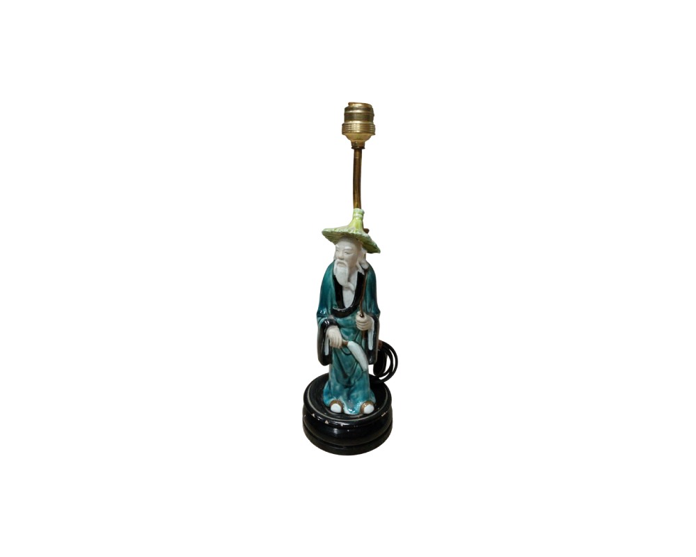 lámpara de sobremesa estructurada a partir de una estatuilla de cerámica esmaltada que representa a un personaje tradicional anciano con túnica azulada y sombrero cónico, montada sobre una base circular oscura y equipada con un vástago y portalámparas metálico superior alquiler para eventos, publicidad, peliculas y series
