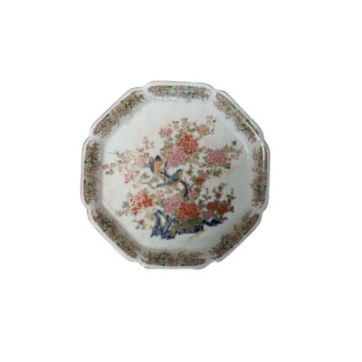A polygonal porcelain plate with lobed edges, characterised by a rich polychrome decoration featuring flowers in shades of pink and orange, birds perched in the centre and a detailed border around the edge; available for hire for events, advertising, films and TV series