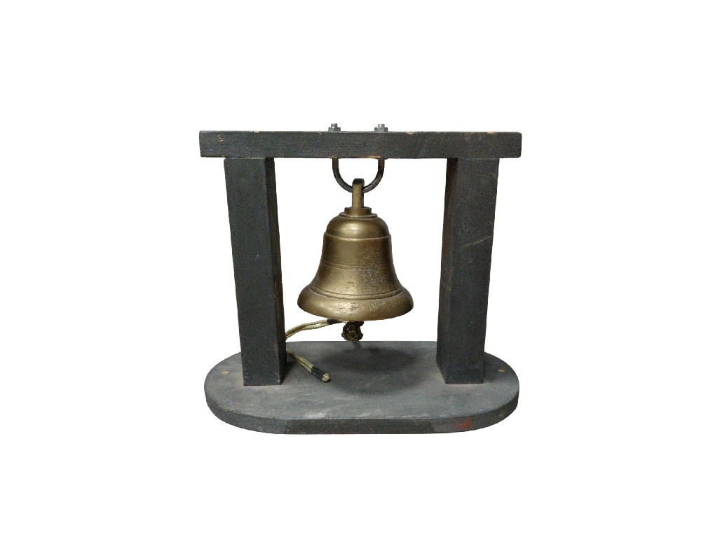 A metal bell (possibly made of brass or bronze) suspended from a simply designed wooden stand. It features a functional clapper to produce a sound and a pull cord. It is a desk or shelf piece that combines a nautical-inspired aesthetic with a decorative or audible signalling function. Available for hire for events, advertising, films and TV series.