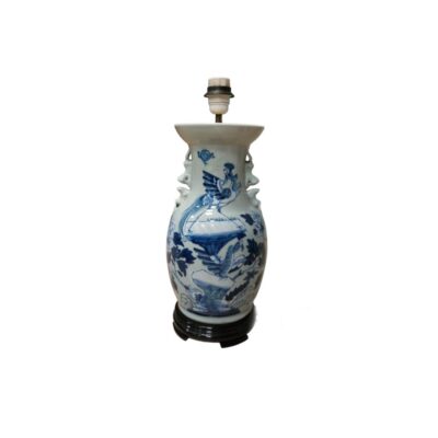 A table lamp crafted from a porcelain vase with blue decoration on a white background, featuring motifs of birds and natural elements. The piece includes a lampshade at the top and rests on a turned wooden base. Available for hire for events, advertising, films and TV series.