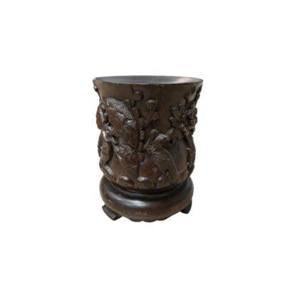 A cylindrical container made of dark wood, finely carved with reliefs depicting natural motifs, such as birds and vegetation. It rests on a circular base with small legs. It is a traditionally styled handcrafted piece, originally designed to hold calligraphy brushes, although it now serves as an elegant decorative item for desks or shelves.