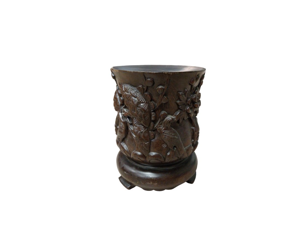 A cylindrical container made of dark wood, finely carved with reliefs depicting natural motifs, such as birds and vegetation. It rests on a circular base with small legs. It is a traditionally styled handcrafted piece, originally designed to hold calligraphy brushes, although it now serves as an elegant decorative item for desks or shelves.