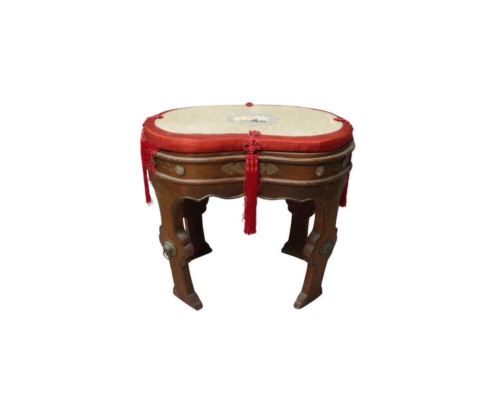 taburete baja de madera con diseño tradicional, caracterizado por patas curvas ornamentadas con apliques metálicos, un asiento superior acolchado en tonos claros y rojos, y detalles colgantes de cordón y flecos rojos alquiler para eventos, publicidad, peliculas y series