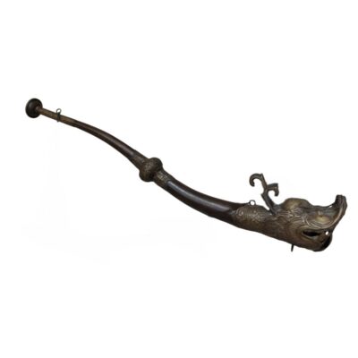 A wind instrument or ornamental piece with an elongated, curved design. It features a structure made of dark metal, topped at one end with a finely sculpted figure depicting the head of a mythical animal (similar to a dragon). The design includes ornamental engravings along its body, lending it an artistic and ceremonial character. It is a handcrafted piece with a traditional aesthetic, ideal as a collector’s item or decorative piece with strong symbolic significance. Available for hire for events, advertising, films and TV series.