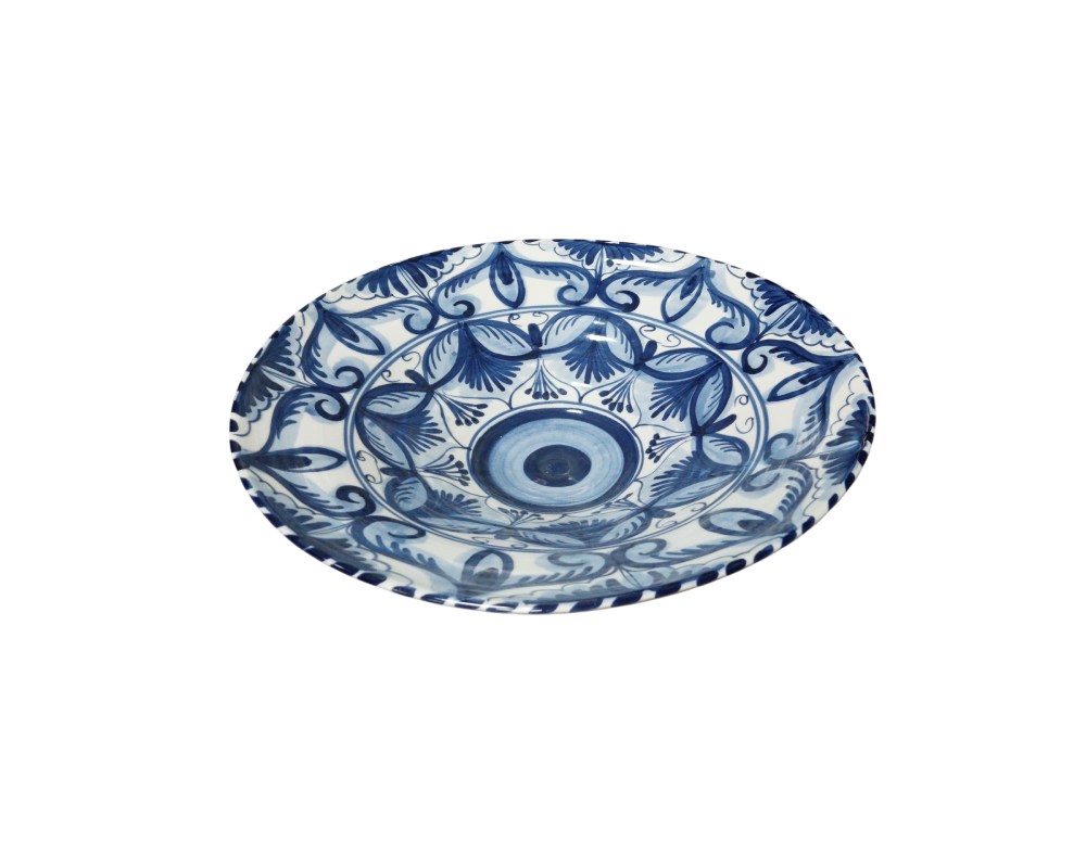 plato decorativo de cerámica que destaca por su fina decoración pintada a mano en azul cobalto sobre un fondo blanco brillante. Su diseño presenta un motivo central circular rodeado por intrincados patrones vegetales y florales estilizados que se extienden hacia el borde, creando una estética simétrica y equilibrada alquiler para eventos, publicidad, peliculas y series