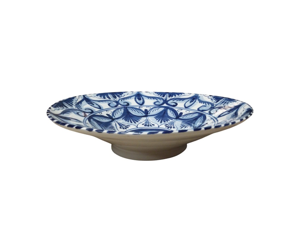 plato decorativo de cerámica que destaca por su fina decoración pintada a mano en azul cobalto sobre un fondo blanco brillante. Su diseño presenta un motivo central circular rodeado por intrincados patrones vegetales y florales estilizados que se extienden hacia el borde, creando una estética simétrica y equilibrada alquiler para eventos, publicidad, peliculas y series