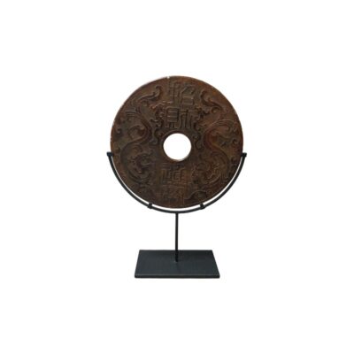 A flat, circular object with a mineral or stony appearance in shades of brown, characterised by a central hole, engravings of traditional characters and intricate reliefs of dragons or sinuous motifs, mounted on a vertical dark metal stand; available for hire for events, advertising, films and TV series