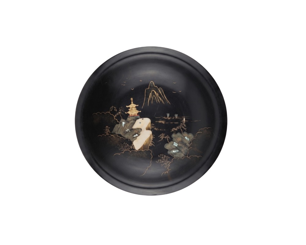 A round, deep plate with a dark surface and a smooth finish, characterised by intricate decoration on the base depicting a traditional landscape featuring mountains, a pagoda and natural elements, highlighted in golden tones and inlays; available for hire for events, advertising, films and TV series