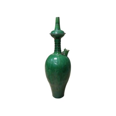 A tall ceramic vessel with a glossy glazed finish in a deep green shade, characterised by a slender body featuring engraved floral reliefs, a zoomorphic spout on the side and a fluted neck culminating in an elaborate, tall, pointed lid; available for hire for events, advertising, films and TV series