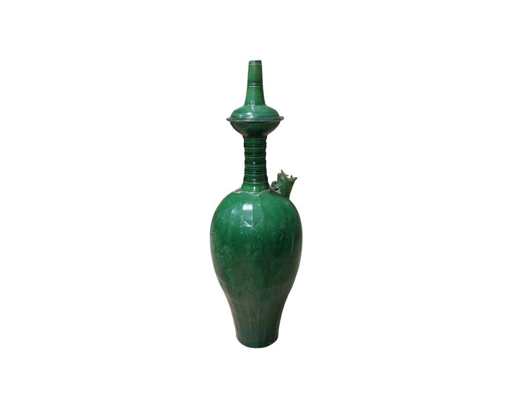 A tall ceramic vessel with a glossy glazed finish in a deep green shade, characterised by a slender body featuring engraved floral reliefs, a zoomorphic spout on the side and a fluted neck culminating in an elaborate, tall, pointed lid; available for hire for events, advertising, films and TV series