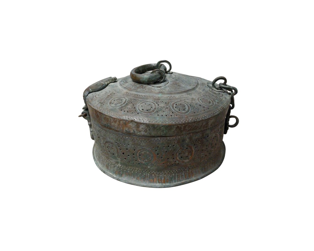 caja de metal de forma circular y aspecto envejecido, caracterizado por una superficie completamente decorada con elaborados grabados y perforaciones de patrones geométricos y florales, una tapa superior con anillas de sujeción y argollas laterales alquiler para eventos, publicidad, peliculas y series