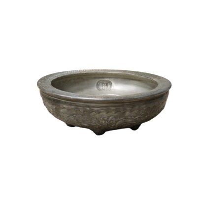 recipiente bajo de cerámica de tonos oscuros y aspecto envejecido, caracterizado por una forma circular con un borde ancho y plano decorado con grabados, un relieve exterior ornamentado alrededor del cuerpo y un pequeño sello o marca distintiva en su interior, apoyado sobre pequeñas patas cortas alquiler para eventos, publicidad, peliculas y series