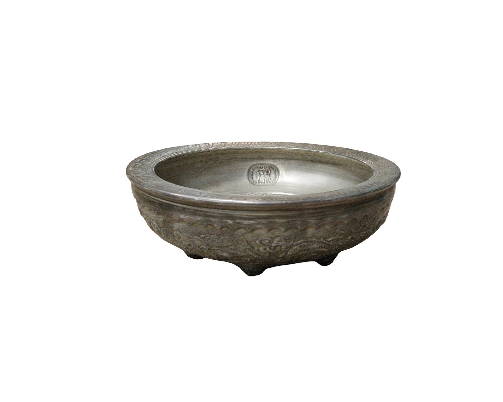 recipiente bajo de cerámica de tonos oscuros y aspecto envejecido, caracterizado por una forma circular con un borde ancho y plano decorado con grabados, un relieve exterior ornamentado alrededor del cuerpo y un pequeño sello o marca distintiva en su interior, apoyado sobre pequeñas patas cortas alquiler para eventos, publicidad, peliculas y series