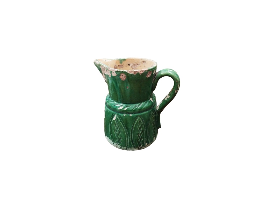 A rustic ceramic milk jug characterised by a cylindrical body decorated with raised motifs on its lower section, a side handle and a pronounced spout; available for hire for events, advertising, films and TV series