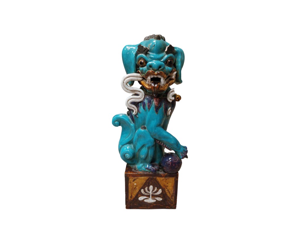 estatuilla de cerámica que representa a un tradicional león guardián o perro de Fu con un llamativo acabado esmaltado en tonos azules y detalles contrastantes, caracterizado por una postura protectora con una pata apoyada sobre una esfera y montado sobre una base cúbica decorada alquiler para eventos, publicidad, peliculas y series