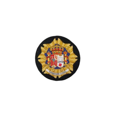 A circular textile emblem, embroidered in gold and coloured threads on a black background, depicting the official coat of arms of Spain and incorporating the institution’s heraldic elements, with a ribbon at the bottom bearing the inscription ‘MINISTERIO FISCAL’. Available for hire for events, advertising, films and TV series.