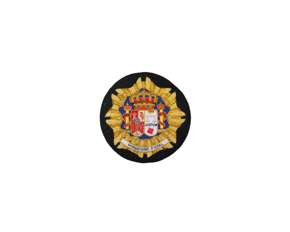 A circular textile emblem, embroidered in gold and coloured threads on a black background, depicting the official coat of arms of Spain and incorporating the institution’s heraldic elements, with a ribbon at the bottom bearing the inscription ‘MINISTERIO FISCAL’. Available for hire for events, advertising, films and TV series.