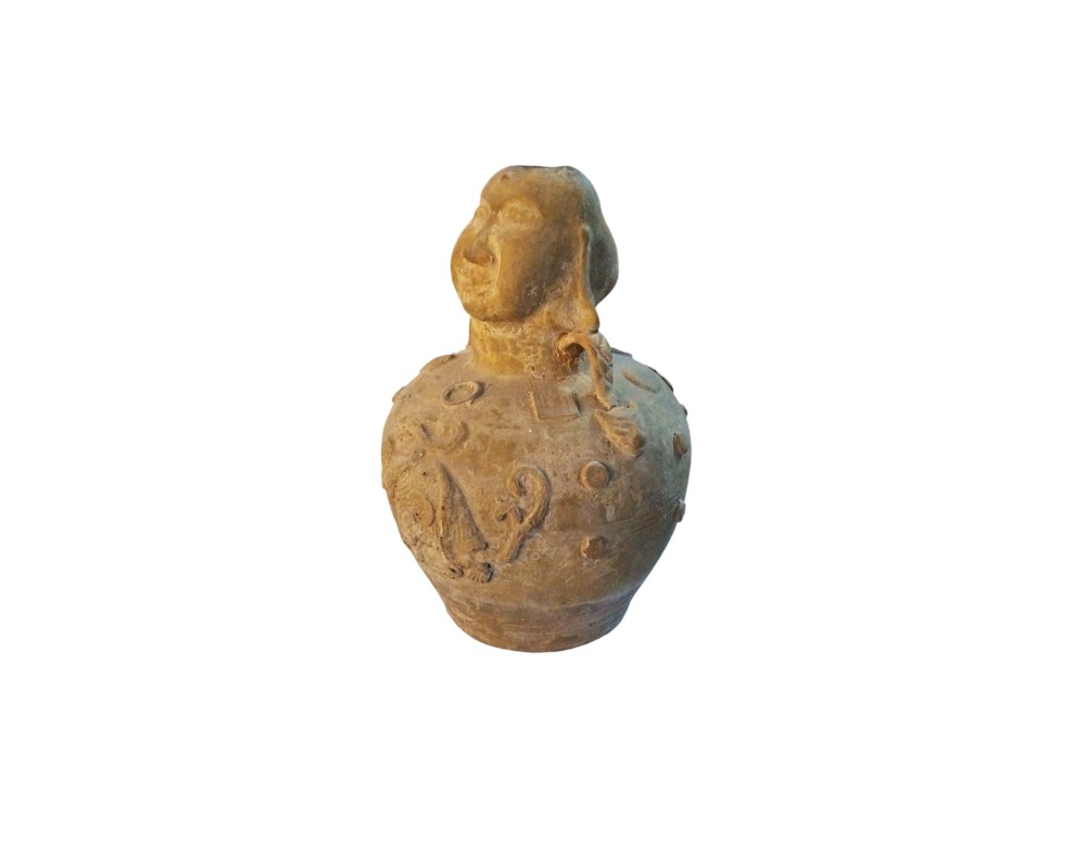 a ceramic vessel in earthy tones with an antique appearance, characterised by a globular body decorated with various reliefs and symbolic motifs distributed across its surface, with a neck at the top sculpted in the shape of a human head or face; available for hire for events, advertising, films and TV series