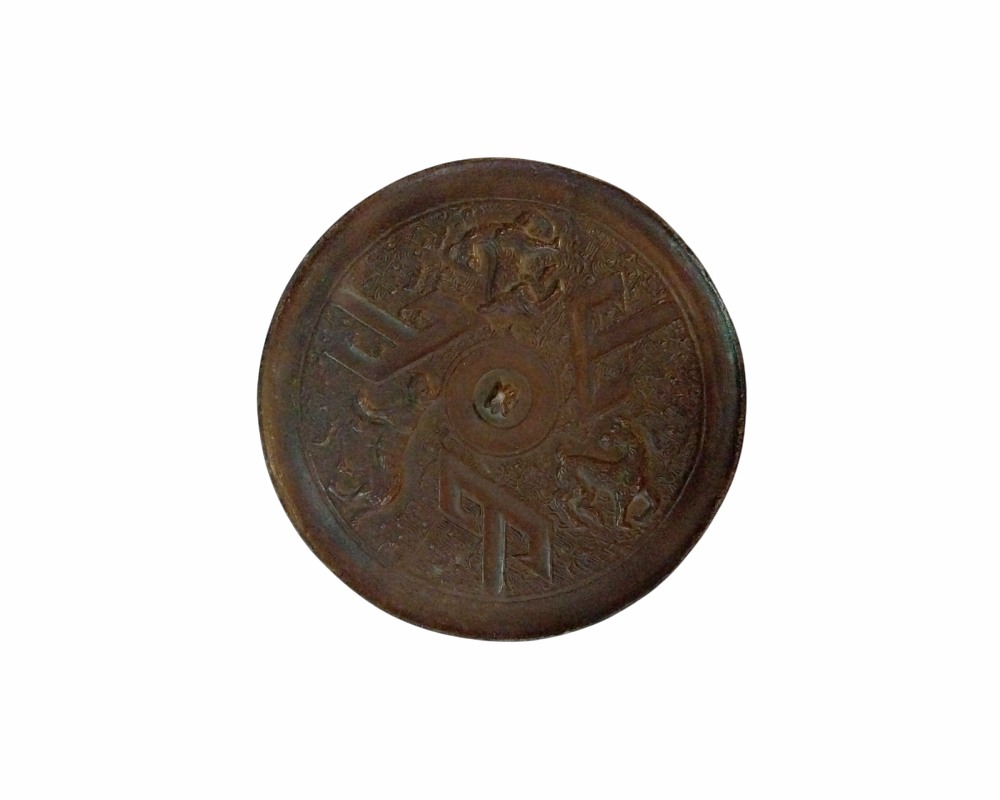 espejo circular de bronce antiguo con un acabado oscuro y envejecido, caracterizado por presentar una superficie ornamentada con un diseño simétrico en relieve que combina elementos geométricos angulares y figuras tradicionales distribuidas en torno a un botón central alquiler para eventos, publicidad, peliculas y series