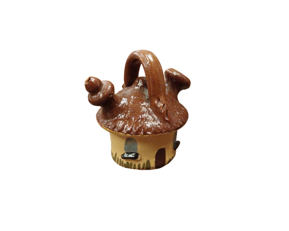 A small ceramic jug modelled in the unique shape of a rustic cottage, featuring a handle at the top, a spout on the side and a glossy brown glaze finish on the top that resembles a roof; available for hire for events, advertising, films and TV series