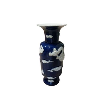 jarrón de porcelana con un acabado esmaltado en tono azul oscuro profundo, caracterizado por un cuerpo cilíndrico, un cuello acampanado y llamativos relieves en color blanco que representan un dragón tradicional entrelazado y motivos de nubes estilizadas a lo largo de su superficie alquiler para eventos, publicidad, peliculas y series