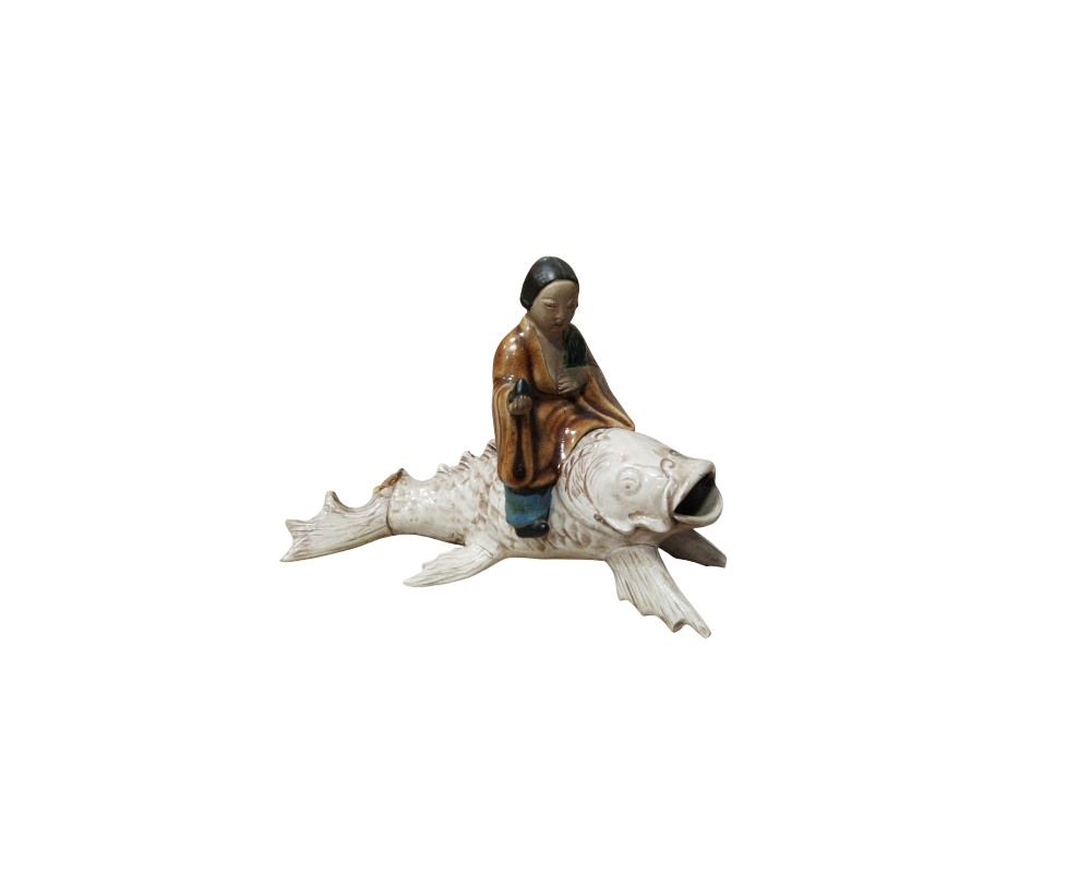 An oriental-inspired decorative ceramic figurine depicting a traditional character dressed in a tunic in shades of brown and ochre, riding on the back of a large, light-coloured fish or carp with detailed fins and an open mouth; available for hire for events, advertising, films and TV series