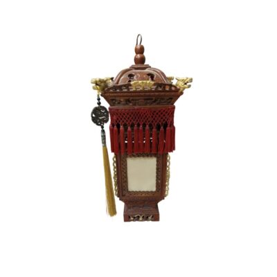 A decorative carved wooden lamp in shades of brown, characterised by an elaborate architectural structure featuring light-coloured panels, embossed gold detailing, decorative dragon heads at the top corners, and a striking fringe design with red tassels and hanging pendants on one side. Available for hire for events, advertising, films and TV series.