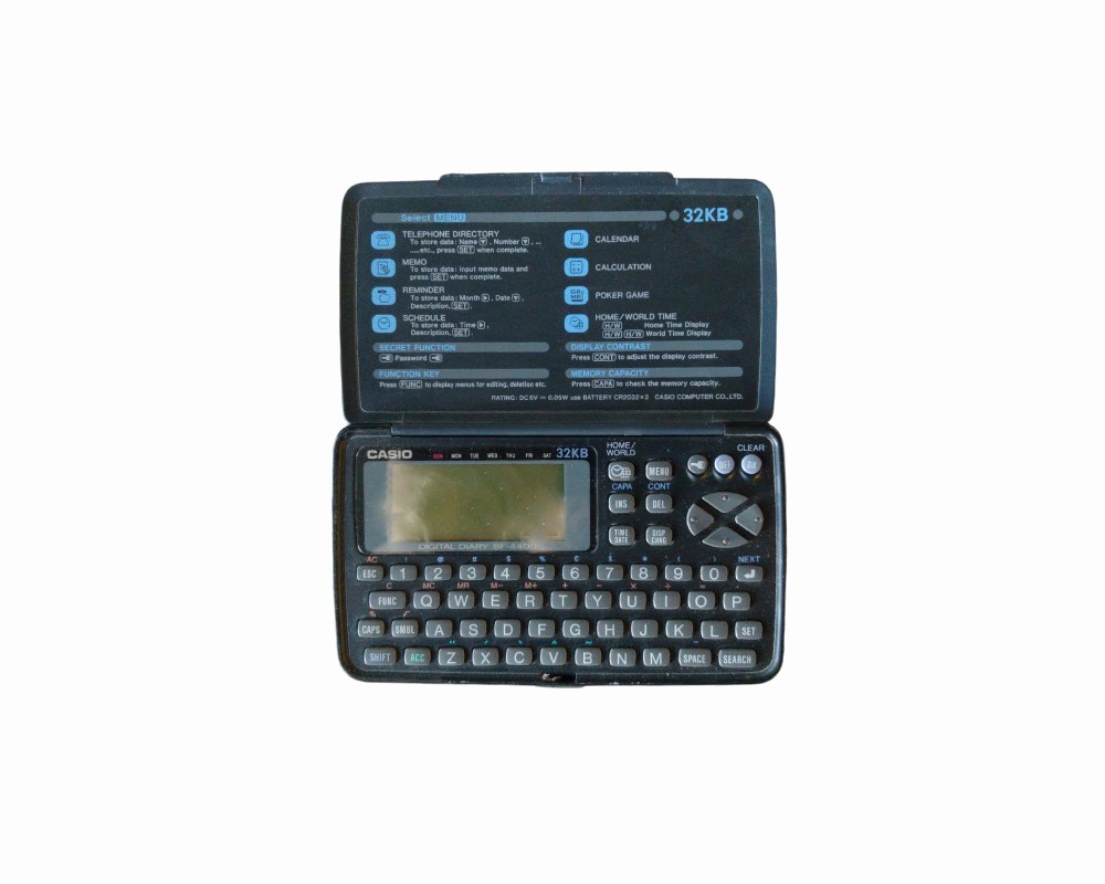 organizador electrónico portátil de la marca Casio, modelo SF-4400 con capacidad de 32KB. Presenta un diseño compacto de concha con carcasa de plástico oscuro, pantalla LCD integrada, teclado completo tipo QWERTY y una cubierta interior que detalla las funciones del menú alquiler para eventos, publicidad, peliculas y series
