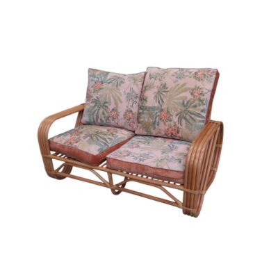 A vintage two-seater sofa crafted from a sturdy frame of bamboo and curved natural rattan, featuring integrated armrests and a set of comfortable separate seat and back cushions upholstered in fabric with a distinctive floral print and tropical-inspired motifs in shades of pink and green. Available for hire for events, advertising, films and TV series.