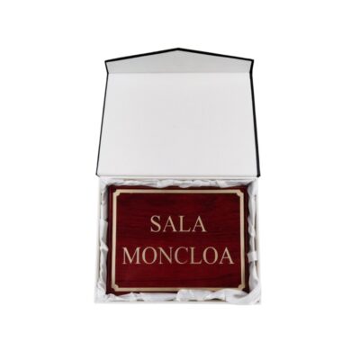 placa rectangular con acabado en madera de tonos rojizos y detalles dorados, grabada con la inscripción "SALA MONCLOA", presentada en el interior de un estuche rígido forrado con tela protectora blanca alquiler para eventos, publicidad, peliculas y series