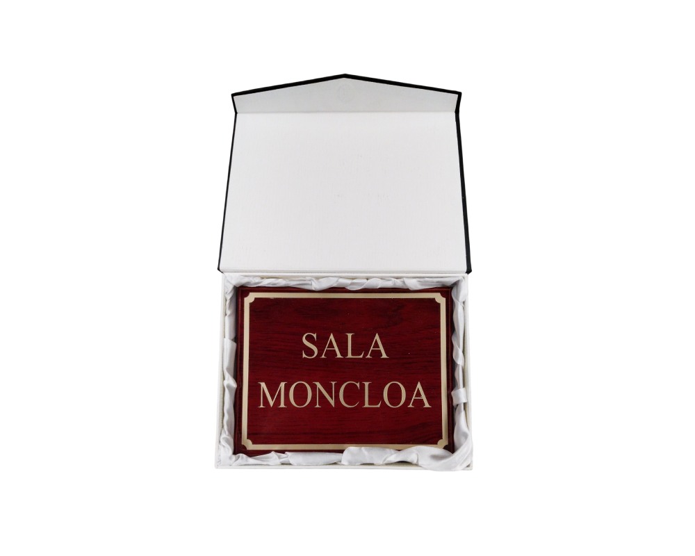 placa rectangular con acabado en madera de tonos rojizos y detalles dorados, grabada con la inscripción "SALA MONCLOA", presentada en el interior de un estuche rígido forrado con tela protectora blanca alquiler para eventos, publicidad, peliculas y series