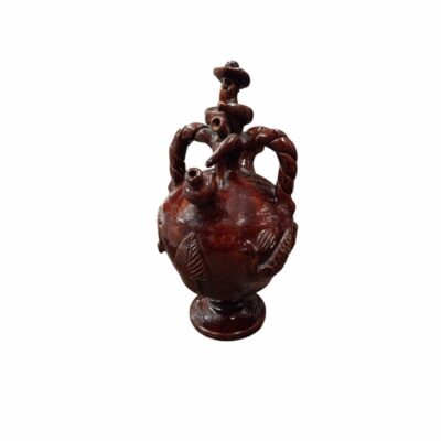 A ceramic jug with a pedestal base and a globular body richly decorated with reliefs of leaves and zoomorphic motifs, featuring two braided side handles, a central spout and a detailed sculpture at the top depicting a traditional figure wearing a hat – available for hire for events, advertising, films and TV series