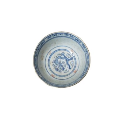 A small ceramic bowl with a glazed finish, featuring delicate cobalt blue decoration depicting a traditional dragon on the inside, surrounded by geometric borders around the rim and subtle reddish inscriptions. Available for hire for events, advertising, films and TV series.