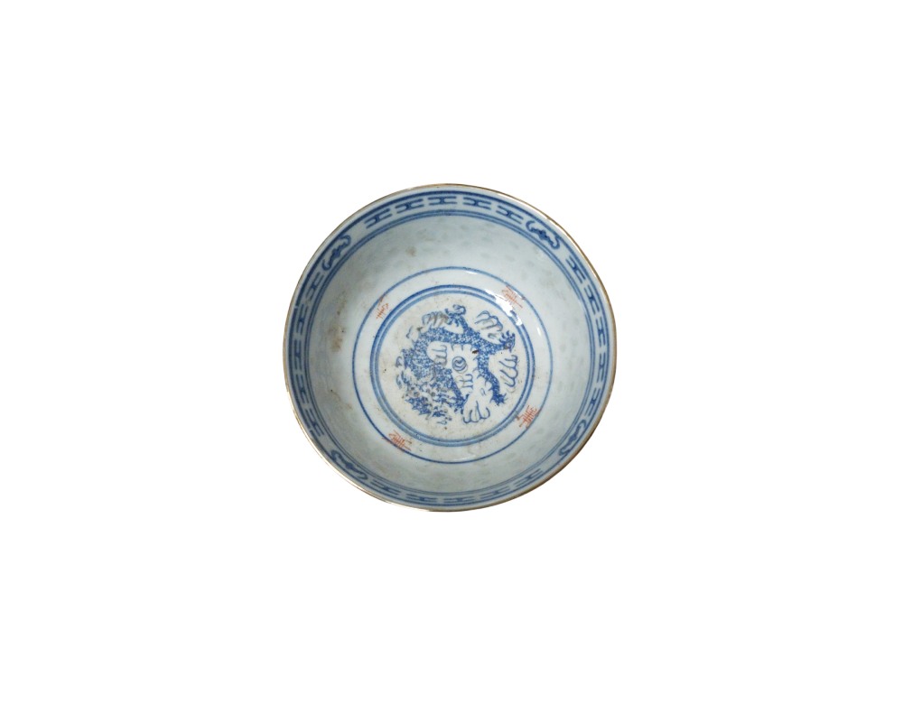 A small ceramic bowl with a glazed finish, featuring delicate cobalt blue decoration depicting a traditional dragon on the inside, surrounded by geometric borders around the rim and subtle reddish inscriptions. Available for hire for events, advertising, films and TV series.