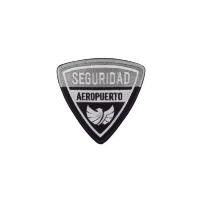 parche textil de forma triangular con bordes bordados, diseñado en tonos grises, blancos y negros, que cuenta con las inscripciones "SEGURIDAD" y "AEROPUERTO", acompañadas de un emblema inferior con silueta de alas alquiler para eventos, publicidad, peliculas y series