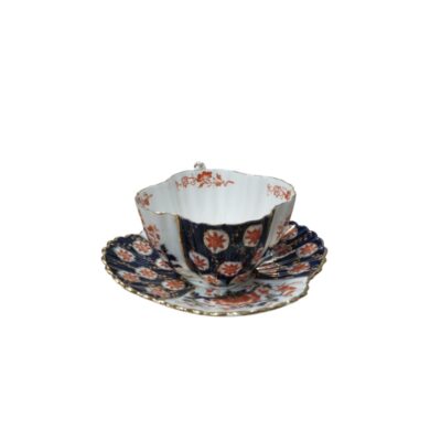 a set comprising a cup and matching saucer made of fine porcelain, characterised by a lobed or wavy silhouette and a rich decoration combining panels in dark cobalt blue, floral motifs in reddish tones and gold-lined details around the rims. Available for hire for events, advertising, films and TV series.