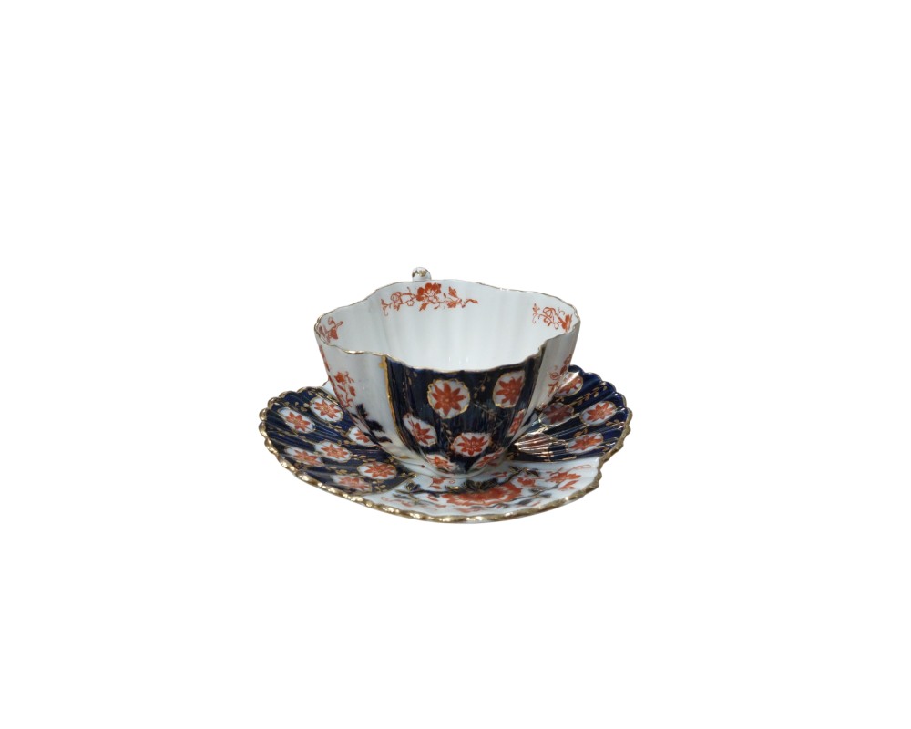 a set comprising a cup and matching saucer made of fine porcelain, characterised by a lobed or wavy silhouette and a rich decoration combining panels in dark cobalt blue, floral motifs in reddish tones and gold-lined details around the rims. Available for hire for events, advertising, films and TV series.