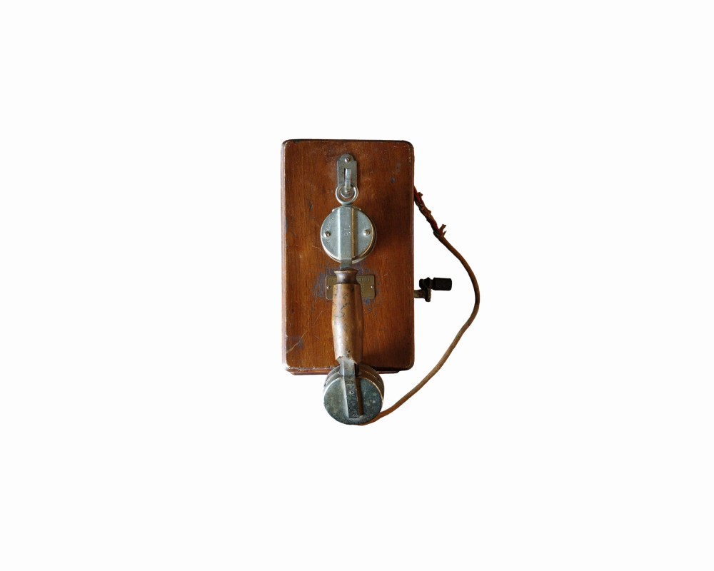 teléfono de pared histórico de época, caracterizado por su sólida carcasa rectangular de madera noble provista de elementos metálicos, manivela lateral y un auricular de tipo monófono con mango de madera central alquiler para eventos, publicidad, peliculas y series