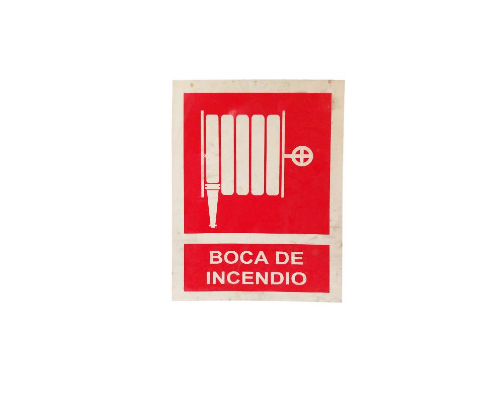 A rectangular, portrait-oriented sign featuring a red panel displaying the white silhouette of a fire hose or hose reel, accompanied by the inscription ‘BOCA DE INCENDIO’ in white lettering at the bottom; available for hire for events, advertising, films and TV series